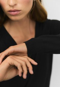 Maglione nero a maglia con scollo a V, con maniche aderenti. Primo piano di mani nude appoggiate l'una sull'altra, mostrando un tono di pelle uniforme.