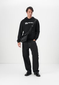 Čierna mikina s kapucňou s logom "KARL LAGERFELD JEANS", skombinovaná s tmavými džínsy a čiernymi topánkami. Na ľavom pleci odpočíva čierny crossbody batoh.