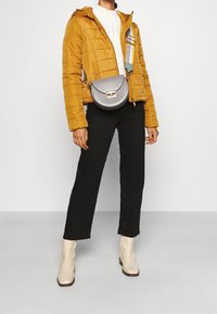 Giacca trapuntata senape, maglione bianco a maglia, pantaloni neri, borsa a tracolla grigia con rifiniture dorate e stivaletti crema. Design e texture semplici.