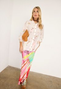 Γυναίκα που φοράει λευκό πουκάμισο με σχέδια και κουμπιά, πολύχρωμη μακριά φούστα, καφέ τσάντα κάτω από το μπράτσο και καφέ παπούτσια με τακούνι, μπροστά σε απλό λευκό τοίχο.