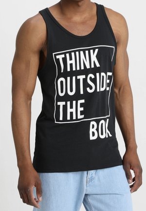 Muskularna osoba ubrana w czarną koszulkę bez rękawów z napisem "THINK OUTSIDE THE BOX" i jasnoniebieskie dżinsy.