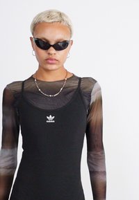 Černé bezrukávové šaty s ribovanou texturou a logem Adidas, vrstvené pod průsvitnou, vzorovanou košilí s dlouhým rukávem, která má gradientní efekt.