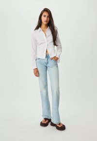 Hvid skjorte med knaplukning, cropped design. Lyseblå utradérende jeans med bleget effekt. Kraftige mørke sandaler. Langt hår stylet nedad, står med hænderne i lommerne.