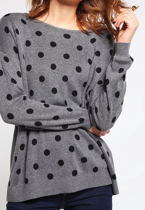 Femme portant un pull gris à manches longues avec des pois noirs, associé à un jean bleu foncé, posant avec une main près de ses cheveux.