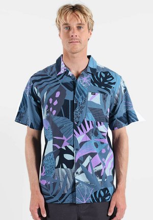 Homme blond portant une chemise à manches courtes à boutons ornée de motifs abstraits de feuilles tropicales en bleu, violet et noir.