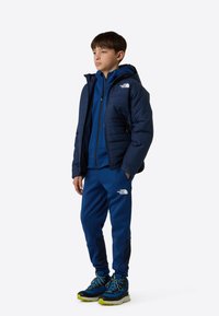 Temno modra puhasta jakna z kapuco, oblečena čez ujemajočo modro jopa z zadrgo in hlače. Črni in modri pohodni čevlji z zelenimi poudarki.