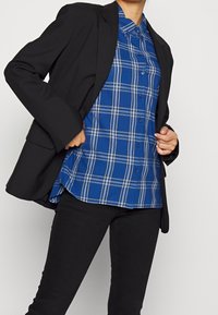 Blazer negro sobre una camisa de cuadros azul con líneas blancas, que cuenta con botones y un cuello puntiagudo. También son visibles unos vaqueros negros.