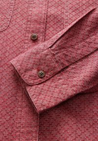Chemise à motifs rouges avec un tissu texturé. Comprend un poignet à boutons et des boutons marron, mettant en valeur un design géométrique subtil sur l'ensemble.