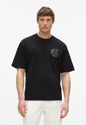 Junger Mann trägt ein schwarzes oversized T-Shirt mit weißen Initialen "KS" auf der linken Brust und weiße Hosen, steht vor einem schlichten Hintergrund.