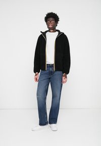 Svart fleece zip-up jakke med gule detaljer, hvit t-skjorte under, blå slitte jeans og hvite joggesko. Står mot en hvit bakgrunn.