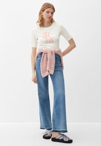 T-shirt blanc à manches courtes avec un motif coloré, un pull en laine rose noué à la taille, un jean en denim bleu à jambe large et des sandales grises avec des semelles noires.