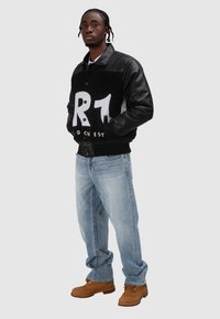 Czarno-biała kurtka bomberka z polaru z skórzanymi rękawami, z dużym nadrukiem i zapięciem na guziki, zestawiona z jasnoniebieskimi dżinsami i brązowymi butami.