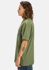 Olivově zelené tričko s krátkým rukávem a kapsou, s černým logem. Vyrobeno z bavlny, má volný střih a hladkou texturu.