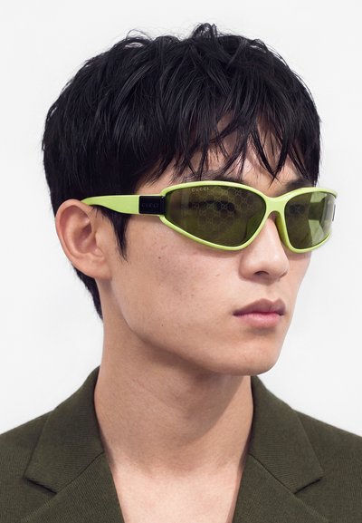 Joven con cabello corto y negro, lleva gafas de sol Gucci con montura verde y una blazer verde oscuro, mirando ligeramente hacia la derecha.
