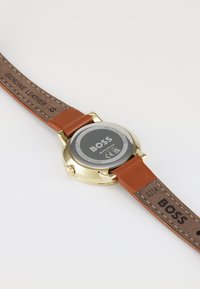 Reloj de tono dorado con una caja redonda de acero inoxidable, correa de cuero genuino marrón y parte trasera grabada que muestra la marca y las especificaciones.
