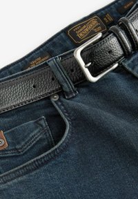 Los pantalones vaqueros de mezclilla azul oscuro presentan un cinturón de cuero negro texturizado con una hebilla de metal plateado, mostrando costuras reforzadas y una apertura en el bolsillo frontal.