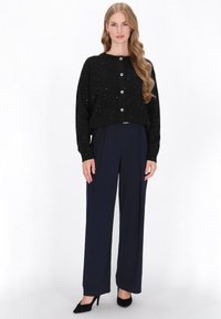 Pulover negru tricotat cu nasturi argintii și detalii ornamentale dispersate, asortat cu pantaloni bleumarin cu talie înaltă și pantofi cu toc înalt negri.