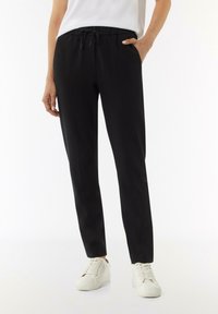 Zwarte, taps toelopende, verstelbare broek gemaakt van een gladde stof; heeft zijzakken en een comfortabele tailleband. Gepaard met witte sneakers.