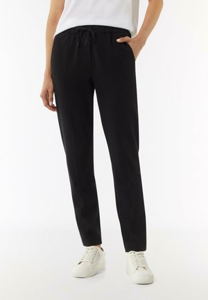 Pantalons noirs, ajustés, à cordon de serrage, fabriqués en tissu lisse ; dotés de poches latérales et d'une taille confortable. Associés à des baskets blanches.