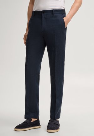 Man draagt marineblauwe op maat gemaakte broek met zijzakken en marineblauwe loafers met gevlochten zolen, staand tegen een lichte achtergrond.