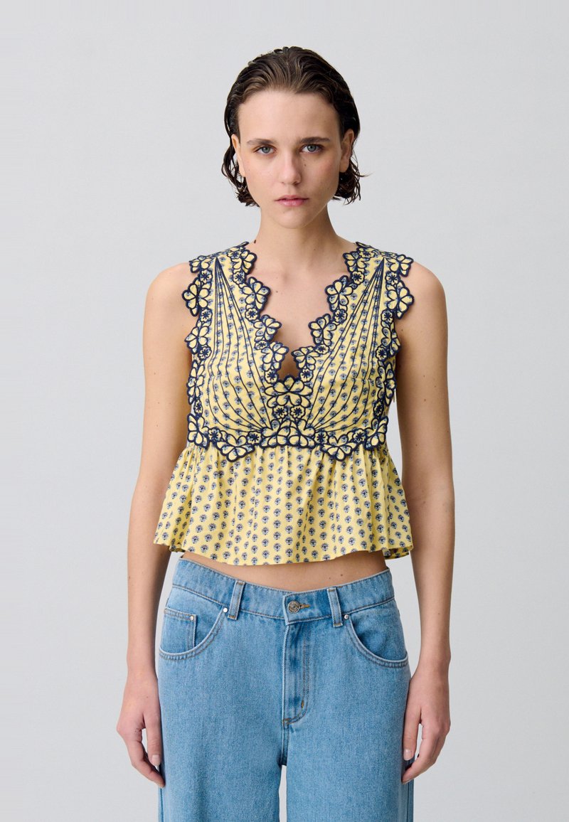 Top giallo a peplum con ricami floreali blu navy, scollatura a V profonda e bordi ruffled. Indossato con jeans a vita alta blu chiaro.