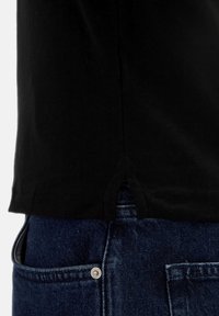 Primo piano dell'orlo di una maglietta nera sopra la tasca di jeans blu con rivetto metallico visibile e dettaglio di spacco laterale.