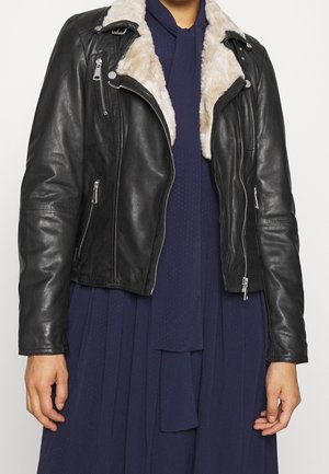 Person iført en sort læder bikerjakke med hvid pelskrave over en mørkeblå kjole med et matchende tørklæde.