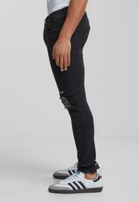 Jeans skinny negros con detalles deshilachados y acentos bordados, combinados con zapatillas blancas y grises con rayas negras y un logo azul.