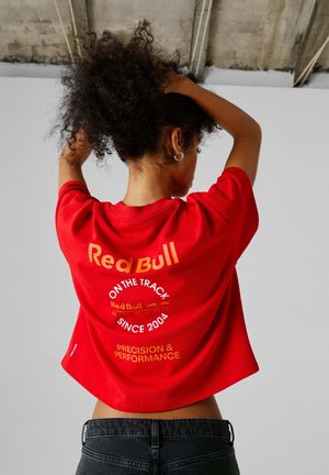Person med krøllete hår iført en rød Red Bull Racing-skjorte som drar håret opp, stående foran en grå vegg og betongtak.