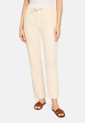 Beige bukser med snøre, båret med brune slip-sandaler, vist fra taljen til fødderne mod en ensfarvet baggrund.