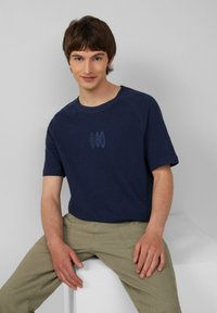 Marineblaue Kurzarm-T-Shirt mit einem runden Halsausschnitt und einer dezenten Grafik von drei hellblauen Formen auf der Vorderseite. Kombiniert mit beigen Hosen.