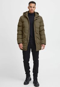 Giacca a vento verde oliva con cappuccio, dotata di chiusura con zip, texture trapuntata e tasche laterali, abbinata a un dolcevita nero e pantaloni neri.
