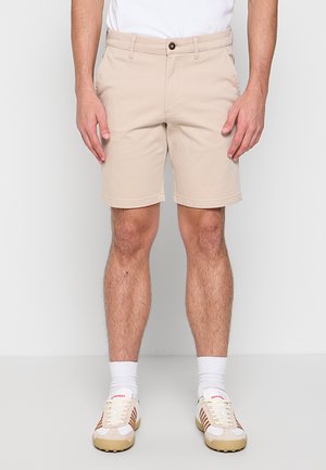 Mann iført beige shorts, hvite ankelsokker og beige og hvite joggesko står mot en ensfarget bakgrunn.