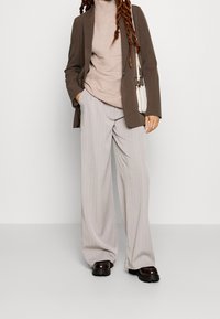 Brązowa marynarka o luźnym kroju, jasnoróżowy sweter z golfem i szerokie spodni w szary paski. Ciemne buty na platformie i mała biała torebka.