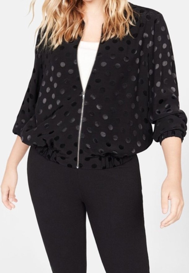 Femme portant une veste noire zippée avec des pois brillants sur un haut blanc et un pantalon noir, avec de longs cheveux blonds ondulés visibles.