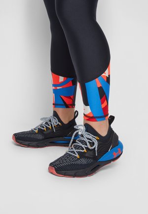 Benen met zwarte legging met rode, blauwe en witte geometrische patronen vlak bij de enkels, gecombineerd met zwarte en blauwe hardloopschoenen met witte veters.