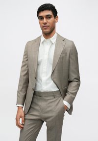 Completo marrone chiaro su misura con una leggera texture, caratterizzato da rever a lancia, due bottoni, abbinato a una camicia bianca con bottoni.