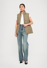 Olijfgroene puffervest, mouwloos met een hoge kraag, voorzien van een ritssluiting en elastiek in de taille, in combinatie met blauwe rechte jeans.