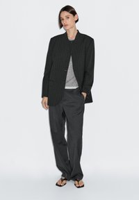 Blazer oversize a righe nere, t-shirt grigia sottostante, pantaloni grigi larghi e ciabatte nere. Tessuto liscio, design minimalista.