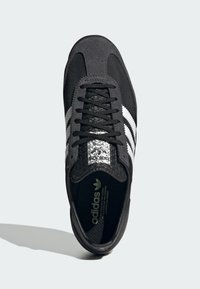Zapatilla negra con un upper de tela suave, diseño de tres rayas plateadas, puntera texturizada, cordones negros y plantilla acolchada.