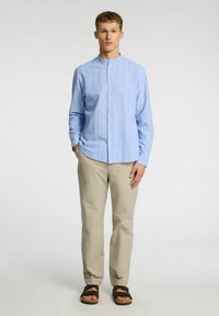 Camisa de botones azul claro con rayas verticales blancas, de manga larga, con cuello mao, combinada con pantalones beiges y sandalias marrones.