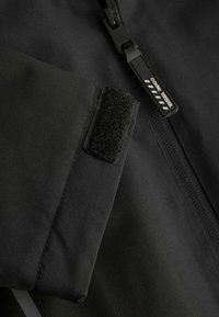 Poignet de veste en tissu noir avec une fermeture auto-agrippante à côté d'un curseur de fermeture éclair noir marqué "code : 1358".