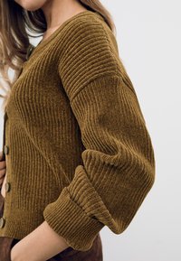 Cardigan marron à côtes avec un tissu texturé. Il dispose d'un col rond, de boutons de fermeture et de manches à revers, mettant en valeur des rayures verticales.