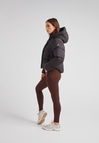 Kobieta w profilu ubrana w czarną kurtkę puchową z kapturem, brązowe legginsy i beżowe buty do biegania na białym tle.