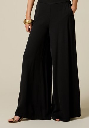 Vrouw draagt zwarte, wijde broek met hoge taille en zijzakken, een grote gouden armband en open teen sandalen met een slangenhuidpatroon.