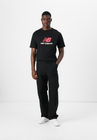 Fekete pamut póló piros "New Balance" logóval, fekete nadrággal párosítva. A modell egy sima fehér háttér előtt áll, fehér sportcipőt visel.