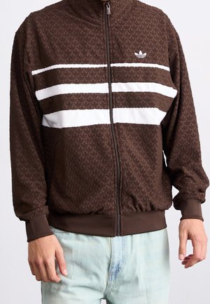 Veste zippée marron texturée avec des rayures blanches horizontales sur la poitrine et un petit logo blanc sur le côté supérieur gauche.