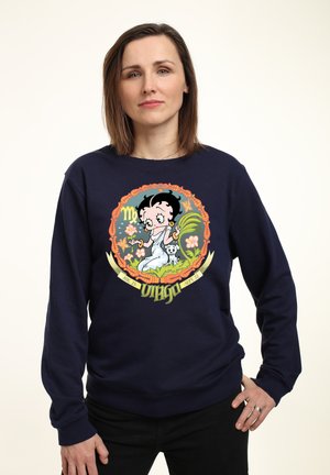 Námorníková mikina s barevným grafickým motivem postavy v bílem oblečení, obklopené rostlinami a květinami, označená "Panna."