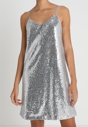 Mujer vestida con un vestido sin mangas plateado con lentejuelas, tirantes finos y escote en V sobre un fondo liso claro.