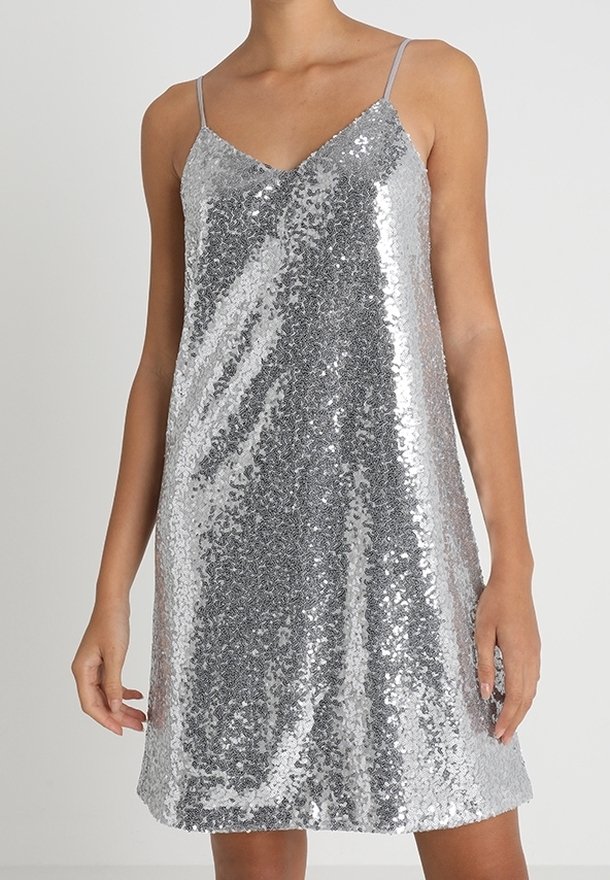 Femme portant une robe sans manches argentée à sequins avec de fines bretelles et un décolleté en V sur un fond clair uni.